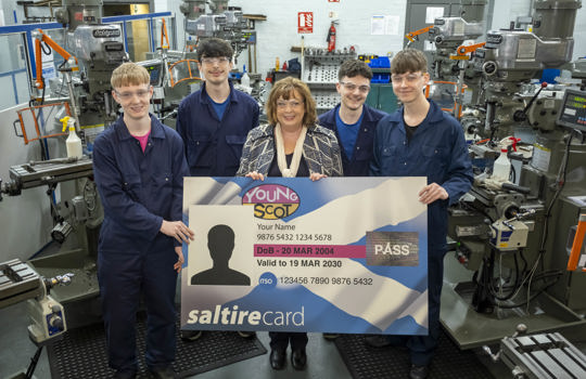 Transport Secretary Fiona Hyslop poses for a photo with a group of young apprentices in a mechanics workshop. The apprentices are wearing overalls and protective glasses. Together, they are holding up a giant version of a young persons' bus pass card.