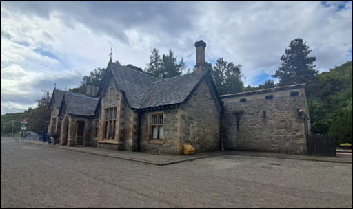 View of Dunkeld and Birnam Station, including Footbridge (Asset 26).  The station is a single-storey high with 7-bays and multiple gables. it is constructed of squared and snecked whinstone rubble with sandstone dressings. It has an open, central porch with pointed arch to front and shouldered arches to sides, advanced gables flanking with stone mullioned tri-partite glazing, decorative barge-boards and pendant timber eaves, tall octagonal and square-cut ridge stacks and a slate roof.